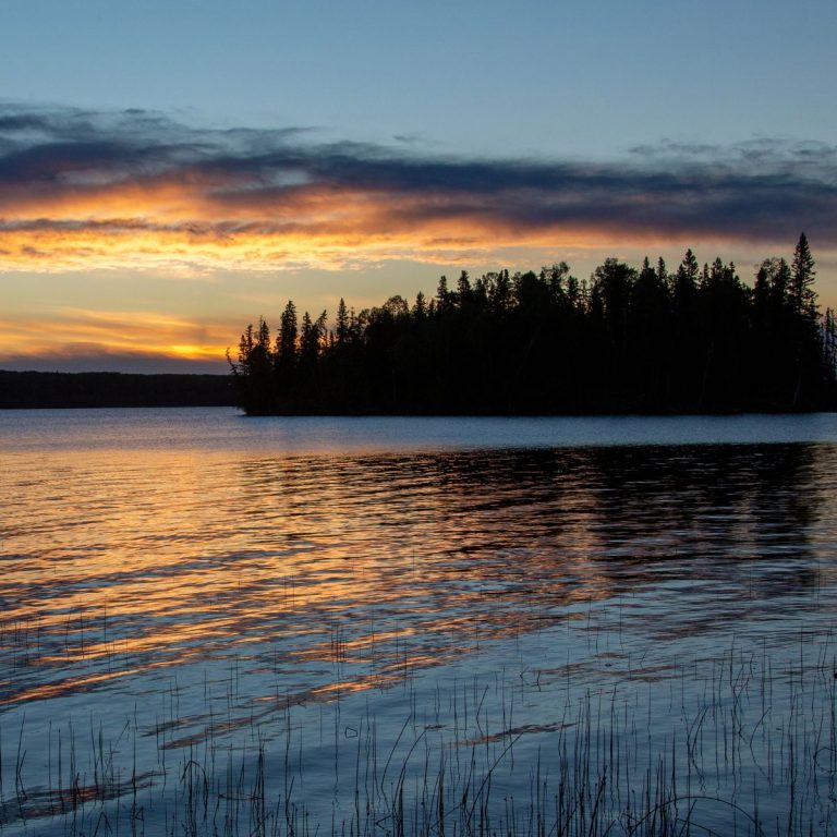 Sunset by the lake, with a fiery sky and some trees in the background.